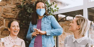 Masked woman stands between two seated unmasked women